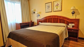 Quarto de hotel com uma cama grande, uma escrivaninha, uma cadeira, duas luminárias fixadas na parede e duas gravuras botânicas emolduradas.
