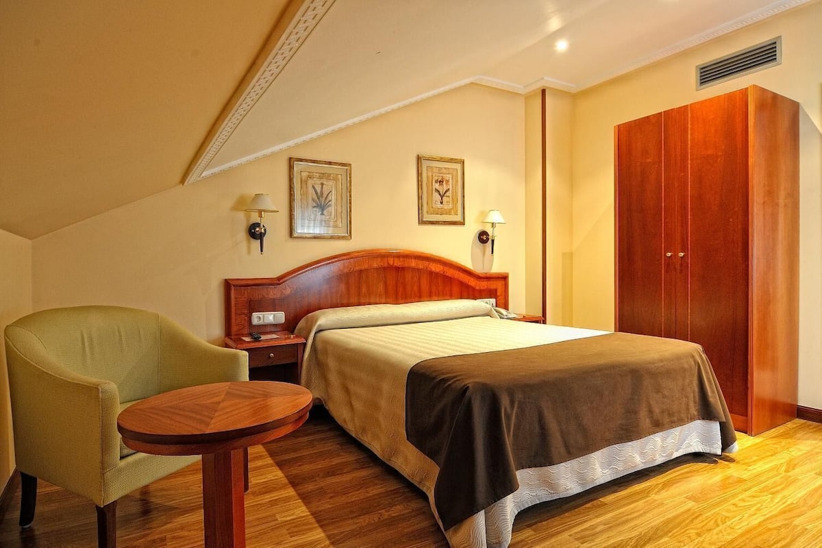 Una habitación de hotel con una cama, una silla, una mesita pequeña, un armario y dos lámparas de pared.