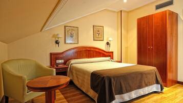 A hotel room with a bed, a chair, a small table, a wardrobe, and two wall-mounted lamps.
