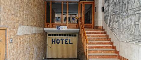 A hotel entrance with a wooden staircase, a marble wall with a relief sculpture, and a 'HOTEL' sign on the door.