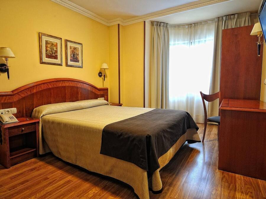 Una habitación de hotel con una cama de madera, dos lámparas de noche, un escritorio con televisor y dos cuadros en la pared.