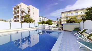 Una piscina con sillones de descanso y un edificio con balcones al fondo.