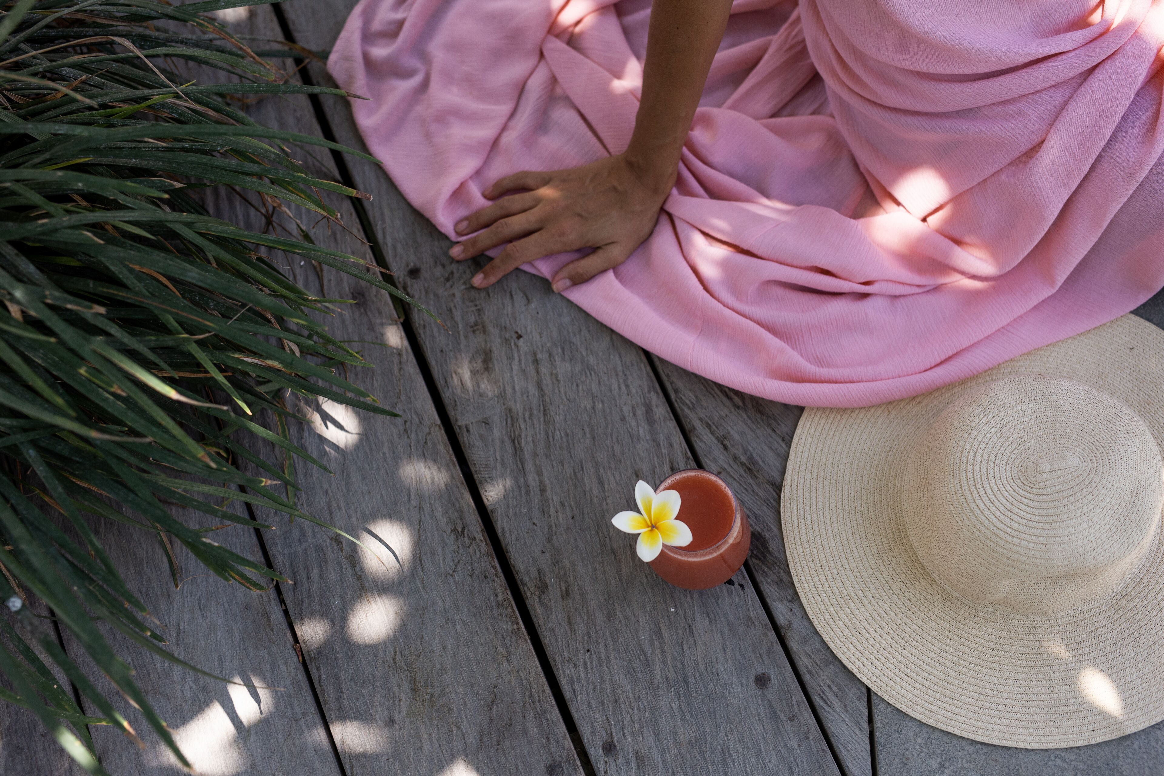 Una terraza de madera con tela rosa, un sombrero de paja y una maceta pequeña con una flor.