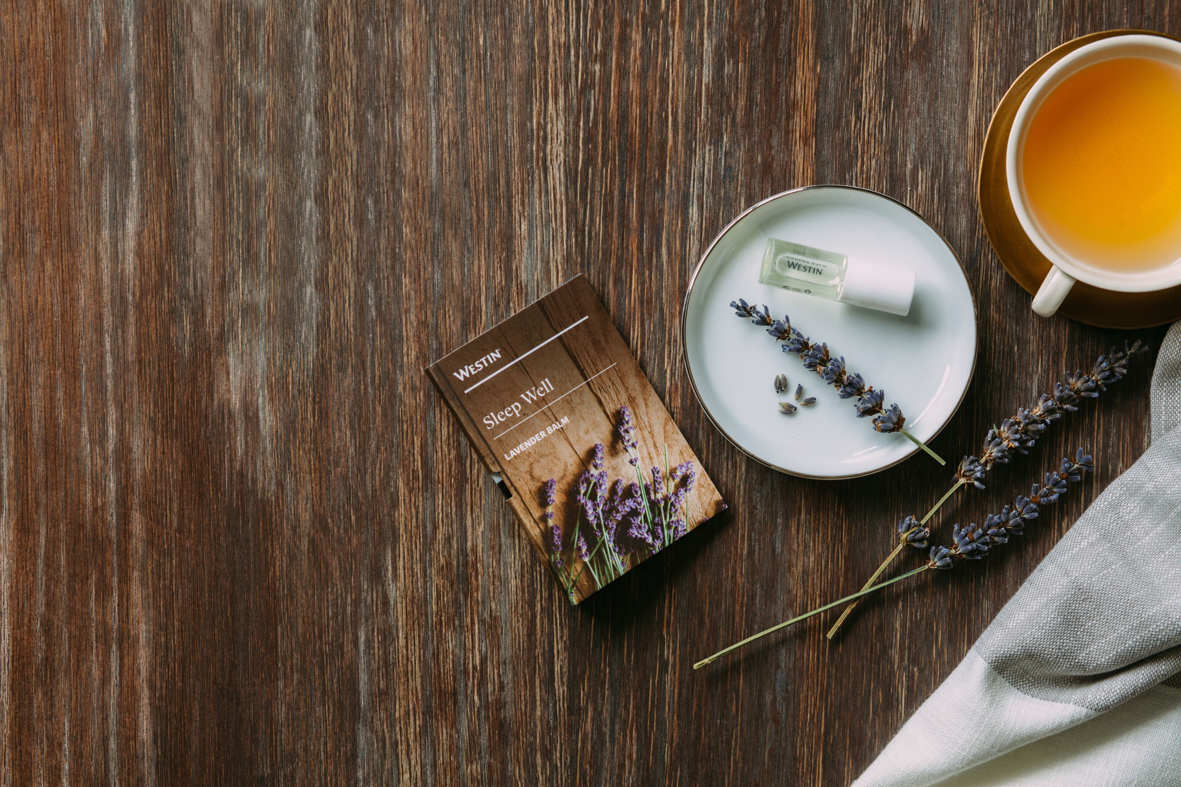 A wooden table with a box labeled "Westin Sleep Well Lavender Balm," a cup of tea, and a lavender sprig.