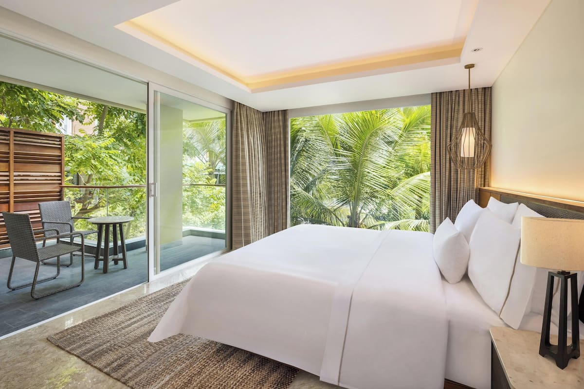 A modern hotel room with a large bed, a balcony with a table and chairs, and a view of lush greenery.