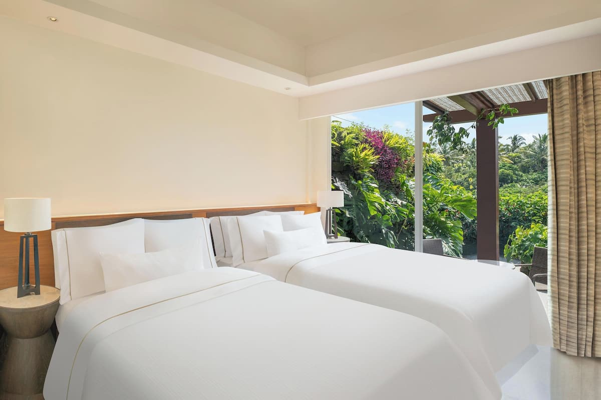 A hotel room with two beds, a nightstand, a lamp, and a large window offering a view of lush greenery.