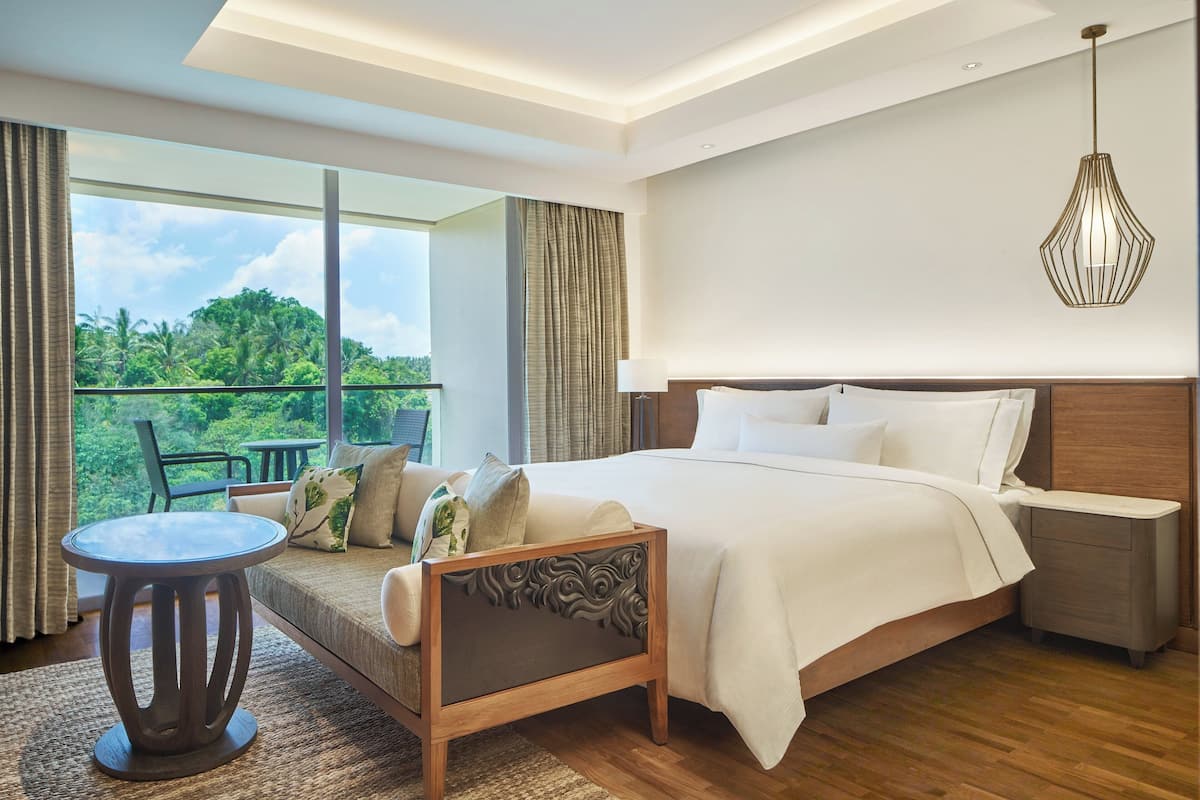 A modern hotel room with a large bed, a small round table, and a view of lush greenery through the window.