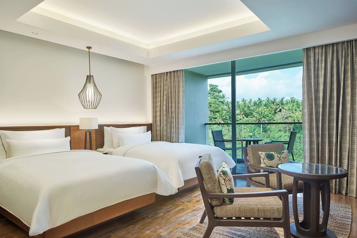 A hotel room with two beds, a sitting area with a table, and a large window with a view of greenery.