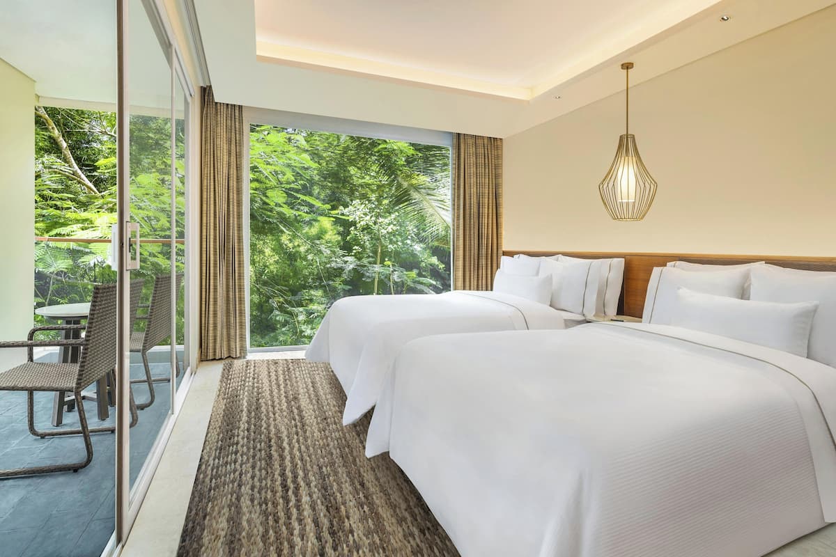 A hotel room with two beds, a large window with a view of greenery, a dining area with chairs, and a hanging light fixture.