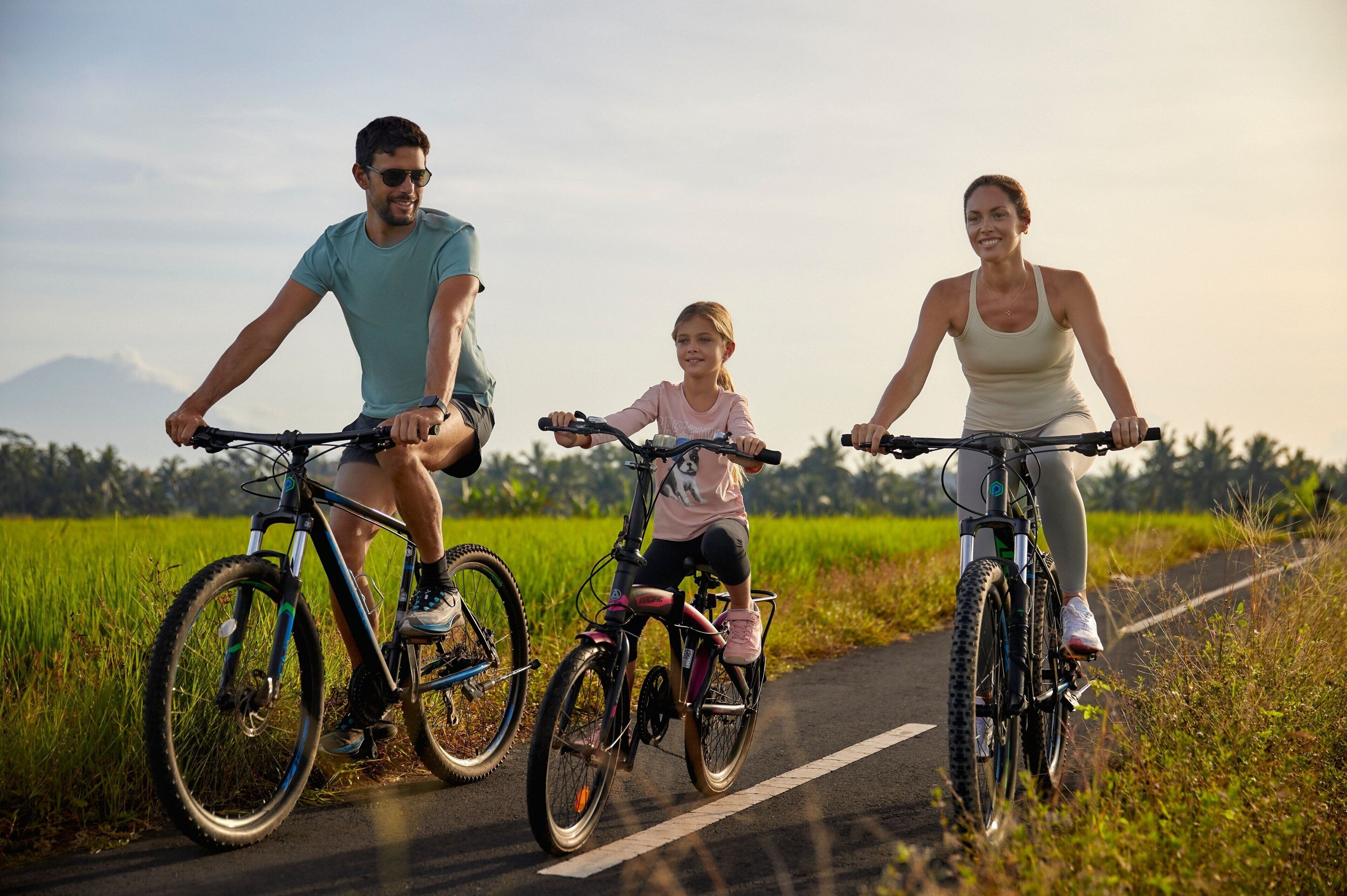 Three people riding bicycles on a road with a green field on one side and a mountain in the background.