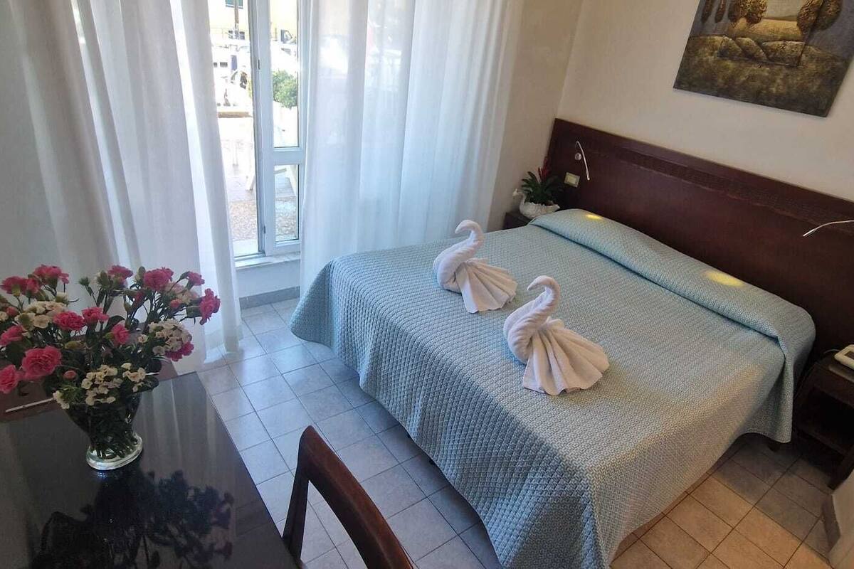 Hotellihuone, jossa on sänky, yöpöytä, maljakko kukkineen ja ikkuna, jossa on verhot.