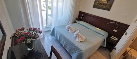 A hotel room with a bed, a bedside table, a vase with flowers, and a window with curtains.