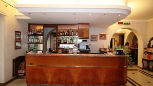 Un hall d’hôtel avec un comptoir d’accueil en bois, un bar avec diverses bouteilles exposées, des tableaux encadrés aux murs et un sol carrelé.
