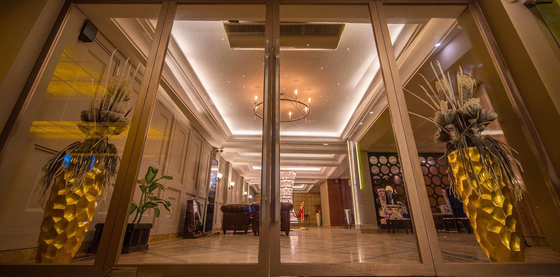 A hotel lobby with a modern design, featuring a central chandelier, decorative plants, and a polished marble floor.