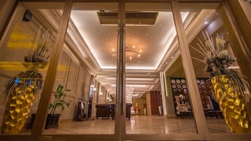 A hotel lobby with a modern design, featuring a central chandelier, decorative plants, and a polished marble floor.
