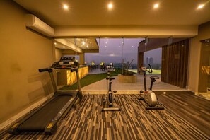 A gym with treadmills and stationary bikes, a glass wall leading to a balcony with a view of a cityscape.