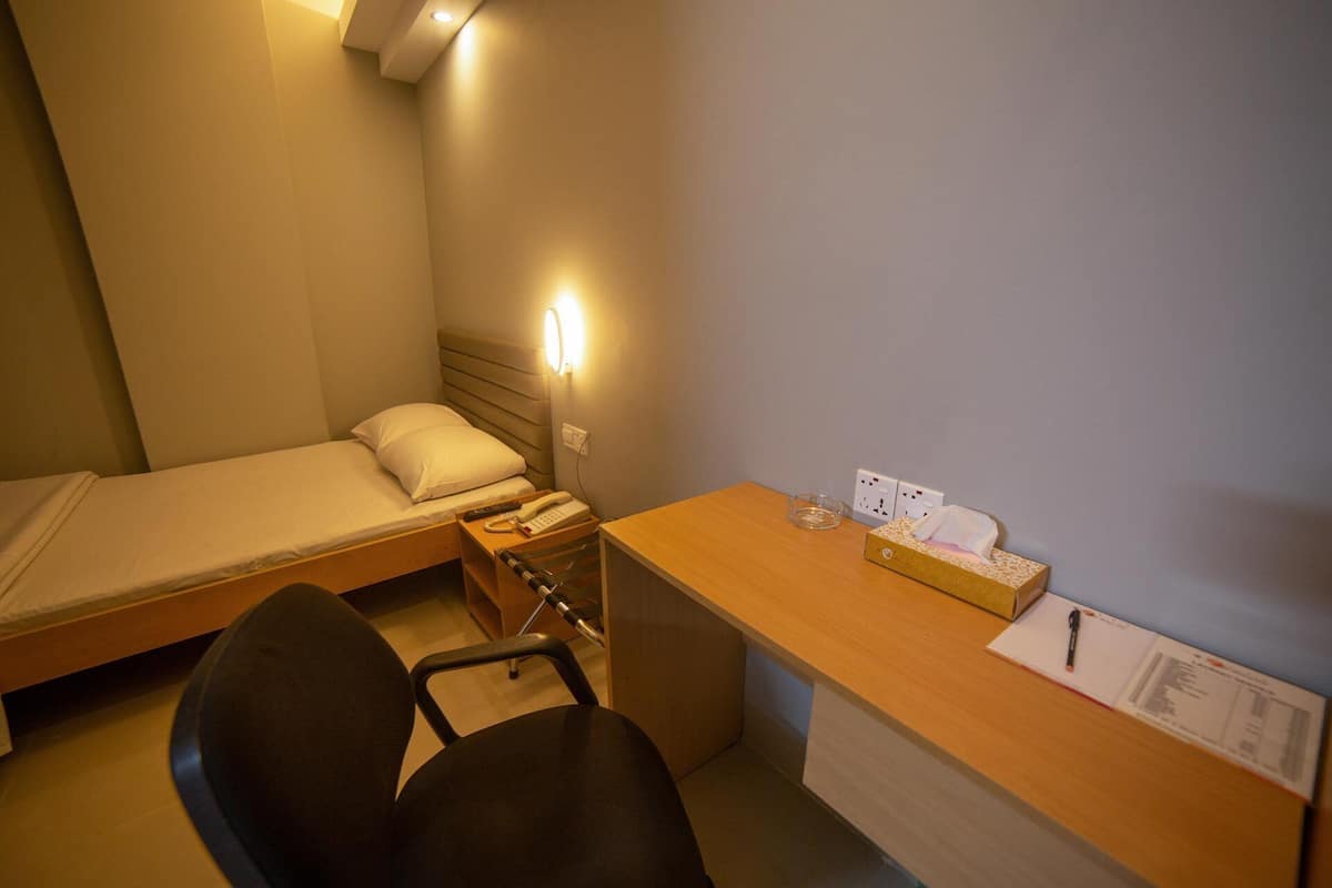 Une petite chambre d’hôtel avec un lit simple, un bureau avec un téléphone, une chaise et une table de chevet avec une boîte de mouchoirs et un stylo.