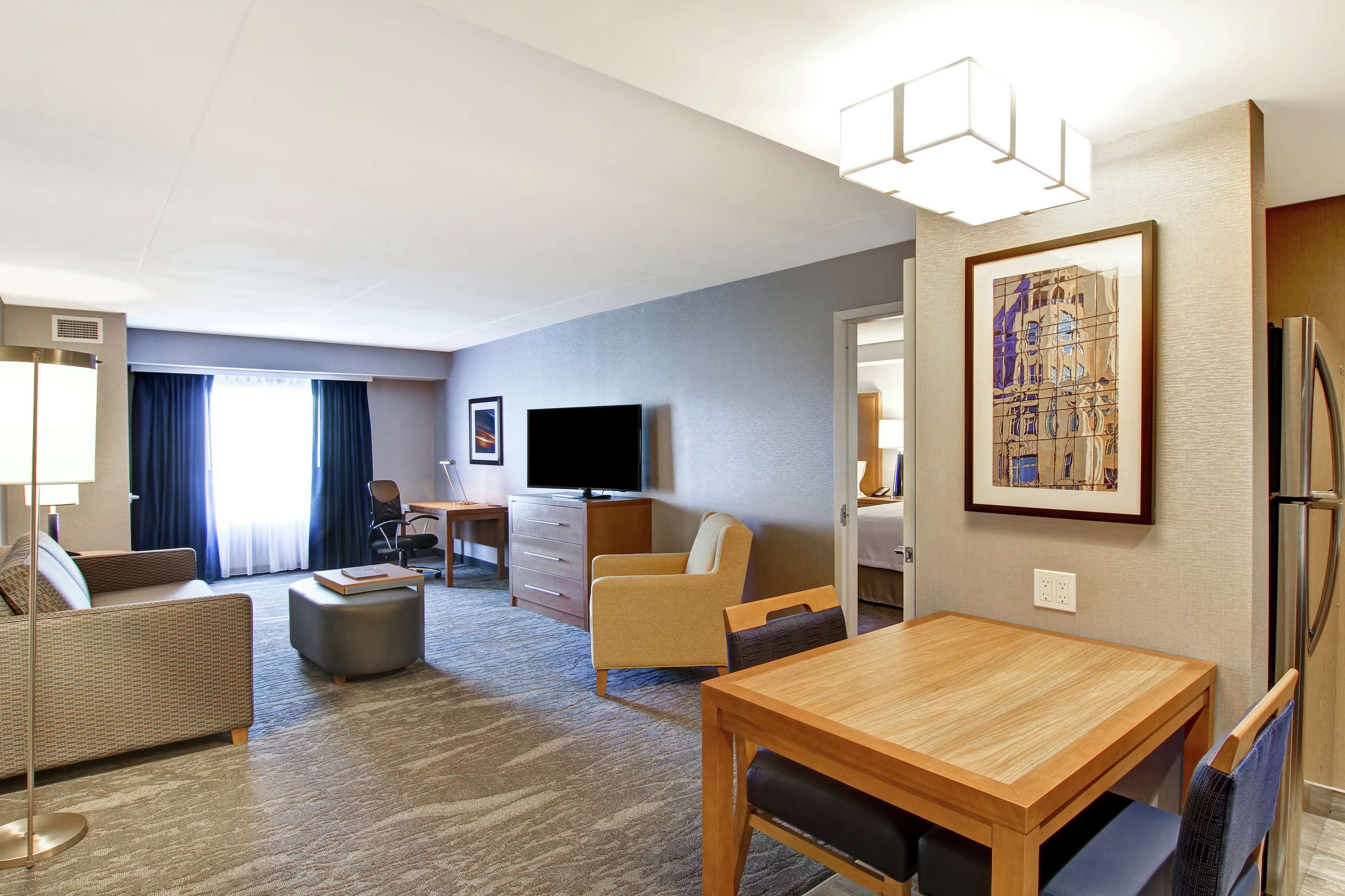 Ein Hotelzimmer mit Wohnbereich, Esstisch, Fernseher, Sofa, Sessel, Schreibtisch und Kühlschrank.