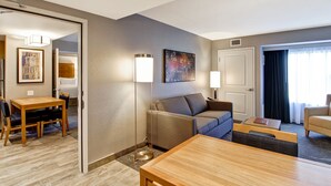Habitación de hotel con zona de comedor, sofá, mesa de centro y un ventanal con cortinas.