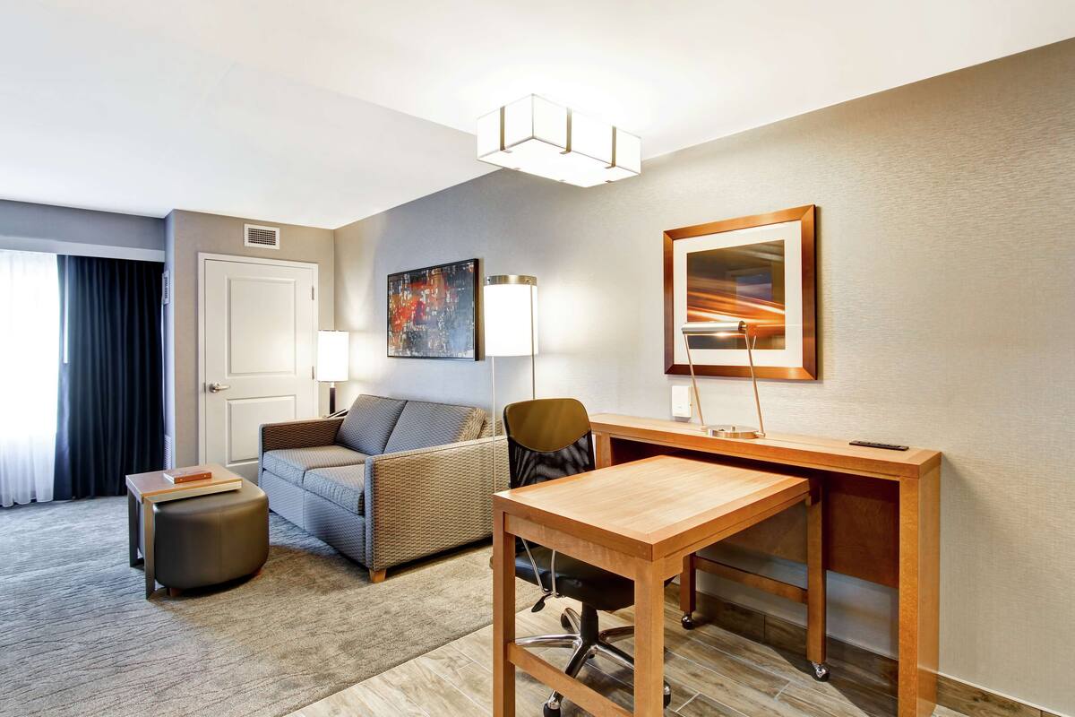 Habitación de hotel con sofá, escritorio, silla, lámpara, un cuadro y ventana con cortinas.