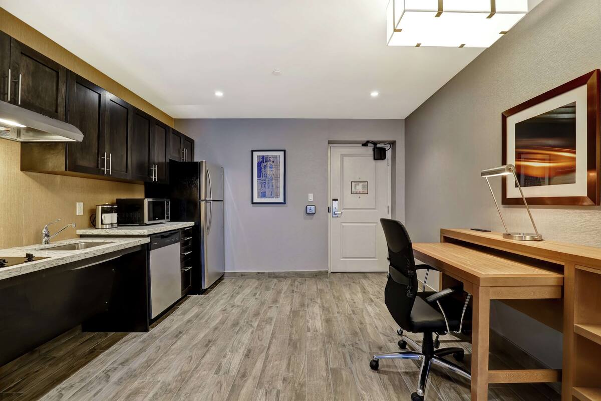 Una cocina moderna con armarios oscuros, un microondas y un lavavajillas. Hay un escritorio de madera con una silla y un cuadro en la pared.