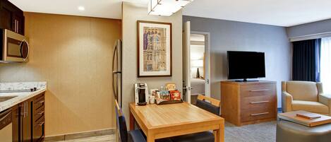 A modern hotel room with a wooden dining table, a sofa, a TV, a coffee machine, and a painting on the wall.