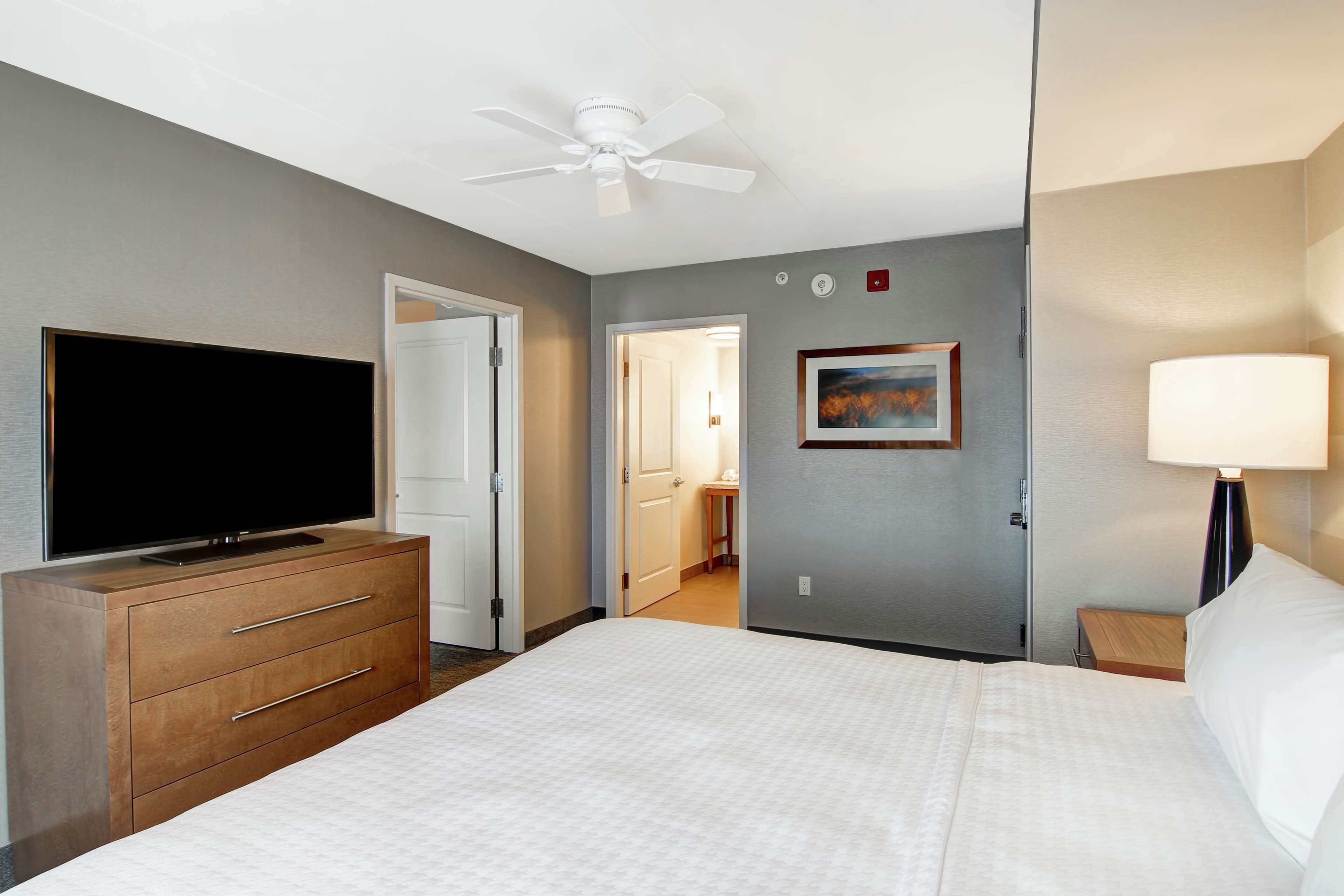 A hotel room with a bed, a TV on a dresser, a ceiling fan, a painting on the wall, and a lamp.