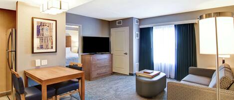 A hotel room with a wooden dining table, a sofa, a TV, and a lamp.