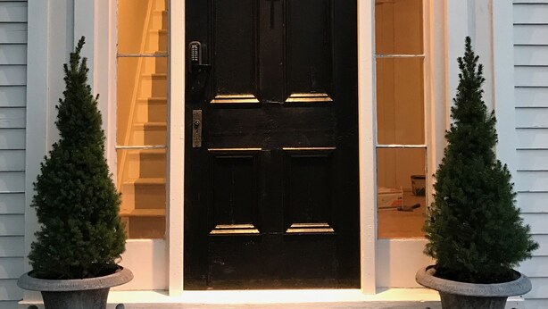 A black front door with a light fixture above it, flanked by two potted evergreen trees. - The Orchard House: Inn & Cafe (Freeport)