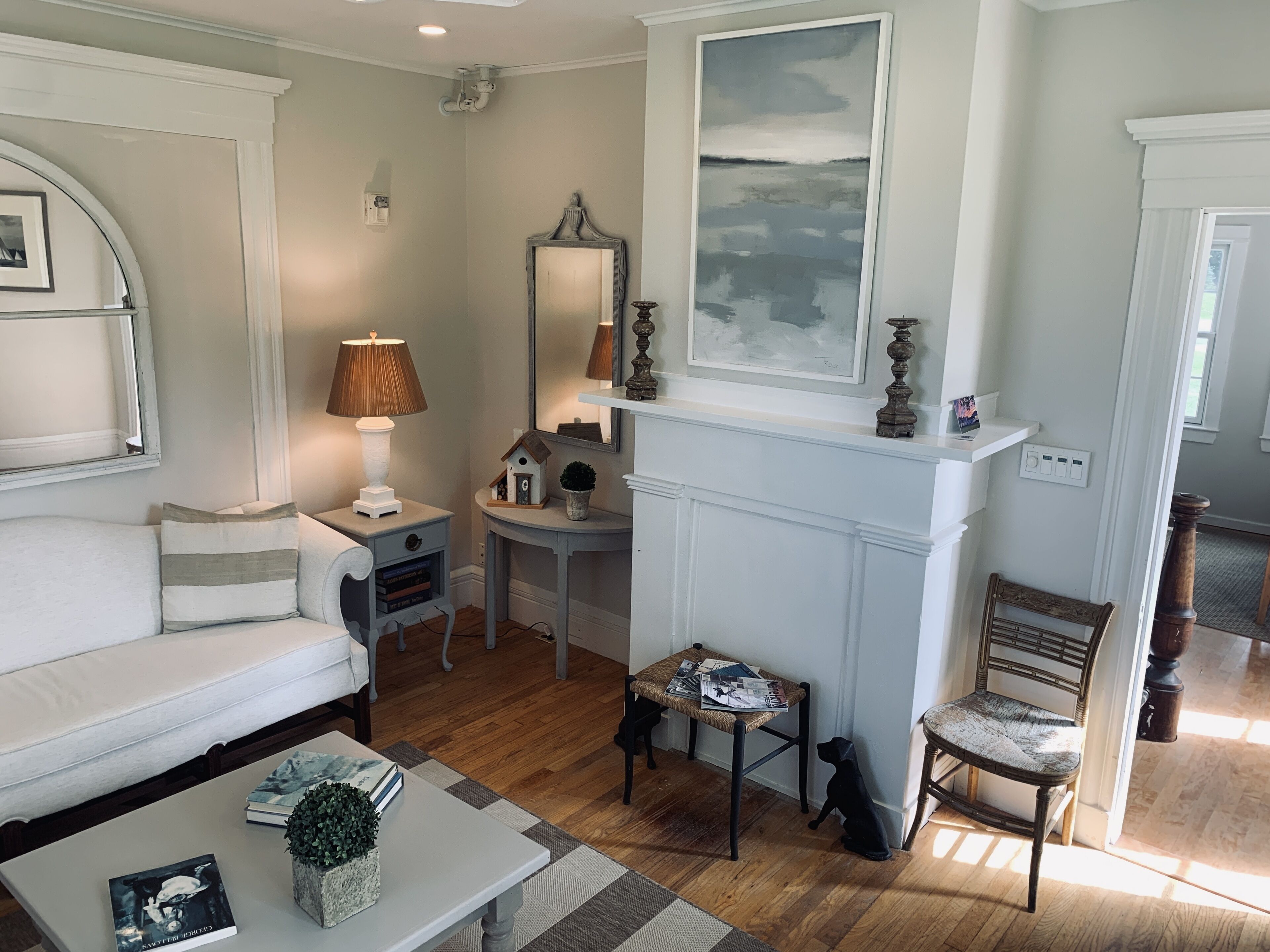 Un salon bien éclairé, avec un canapé blanc, une table basse sur laquelle sont posés des livres, une lampe, une cheminée surmontée d’un tableau et une chaise en bois.