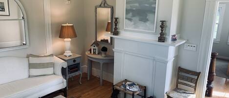 Un salon bien éclairé, avec un canapé blanc, une table basse sur laquelle sont posés des livres, une lampe, une cheminée surmontée d’un tableau et une chaise en bois.