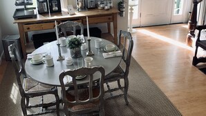 A dining area with a round table set for six, surrounded by wooden chairs. A chandelier hangs above, and a white door with an exit sign is visible in the background. - The Orchard House: Inn & Cafe (Freeport)
