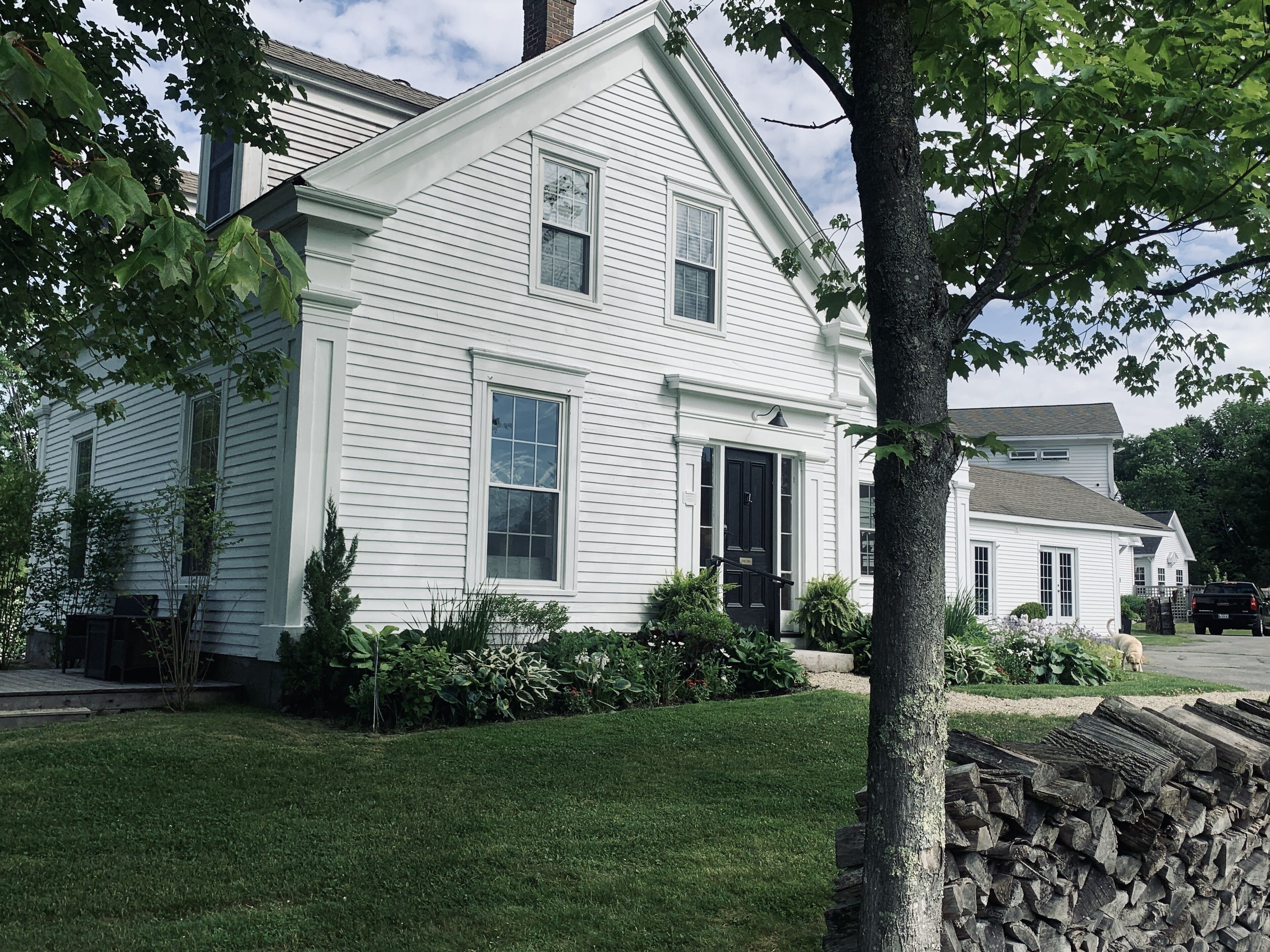 A white two-story house with a front porch, a black door, and a small front yard with a tree and a stack of firewood.