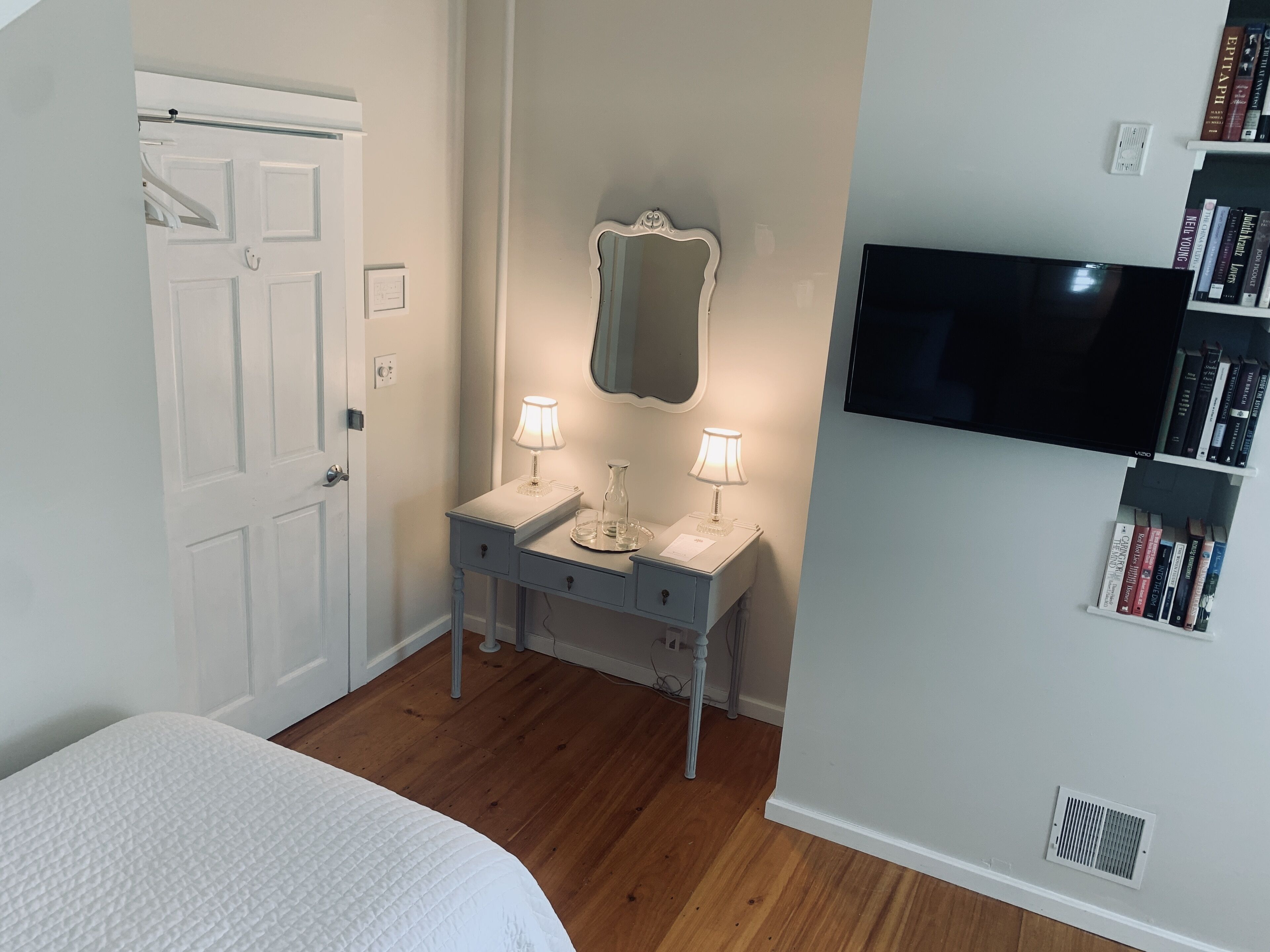 Une chambre à coucher comprenant un lit, une table de chevet avec deux lampes, un miroir, un téléviseur fixé au mur et une bibliothèque.