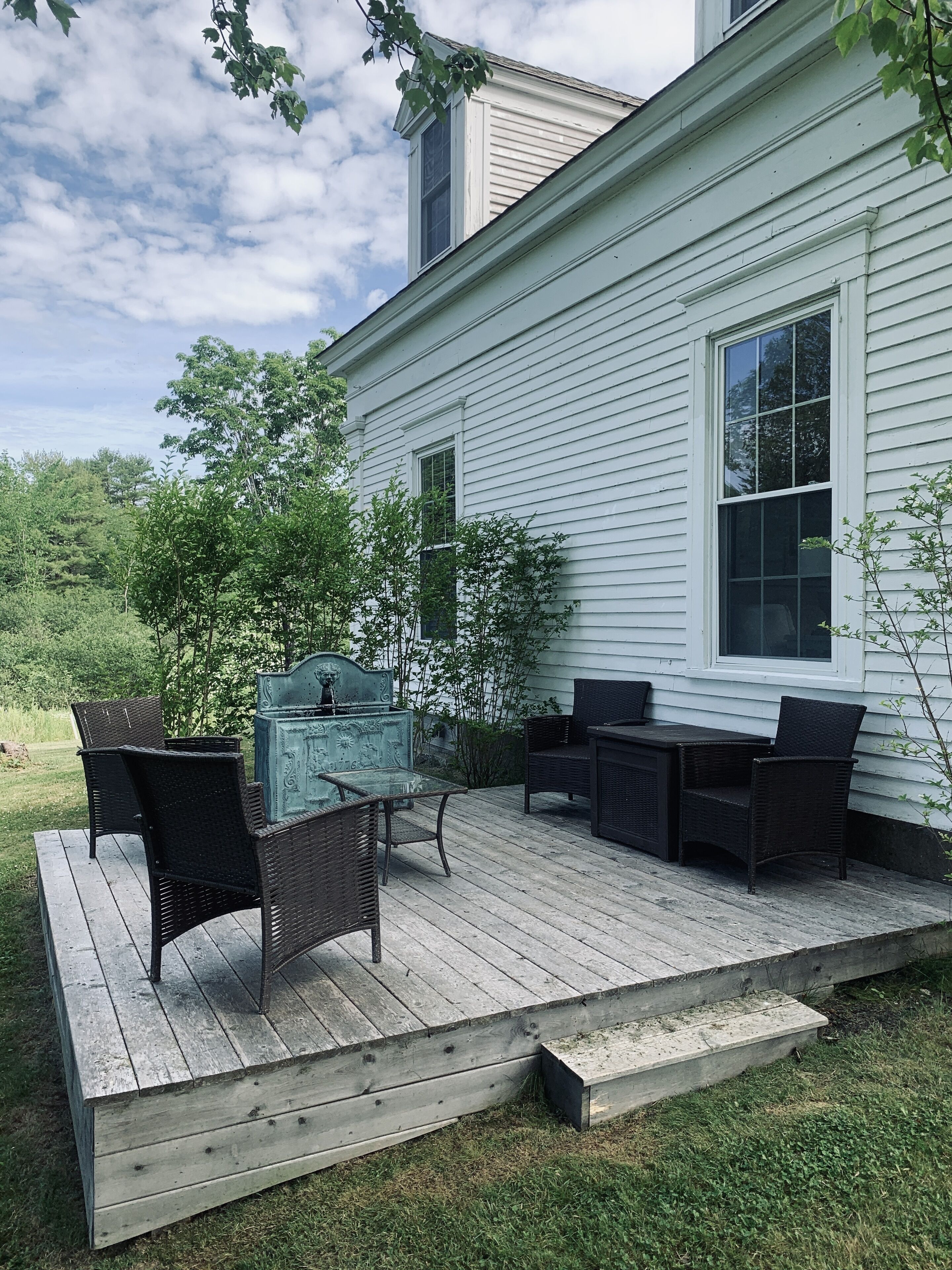 Una terraza de madera con mobiliario exterior, una fuente decorativa y una casa blanca al fondo.