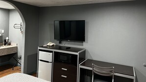 A compact hotel room with a kitchenette, a flat-screen TV mounted on the wall, a small refrigerator, a microwave, and a single chair.