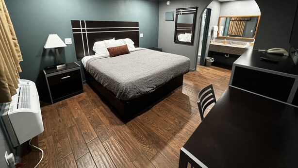 A modern hotel room with a large bed, bedside table with a lamp, a desk with a chair, and a mirror.