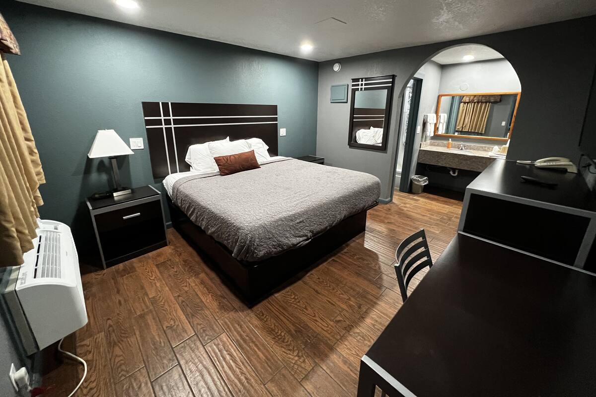 A modern hotel room with a large bed, bedside table with a lamp, a desk with a chair, and a mirror.