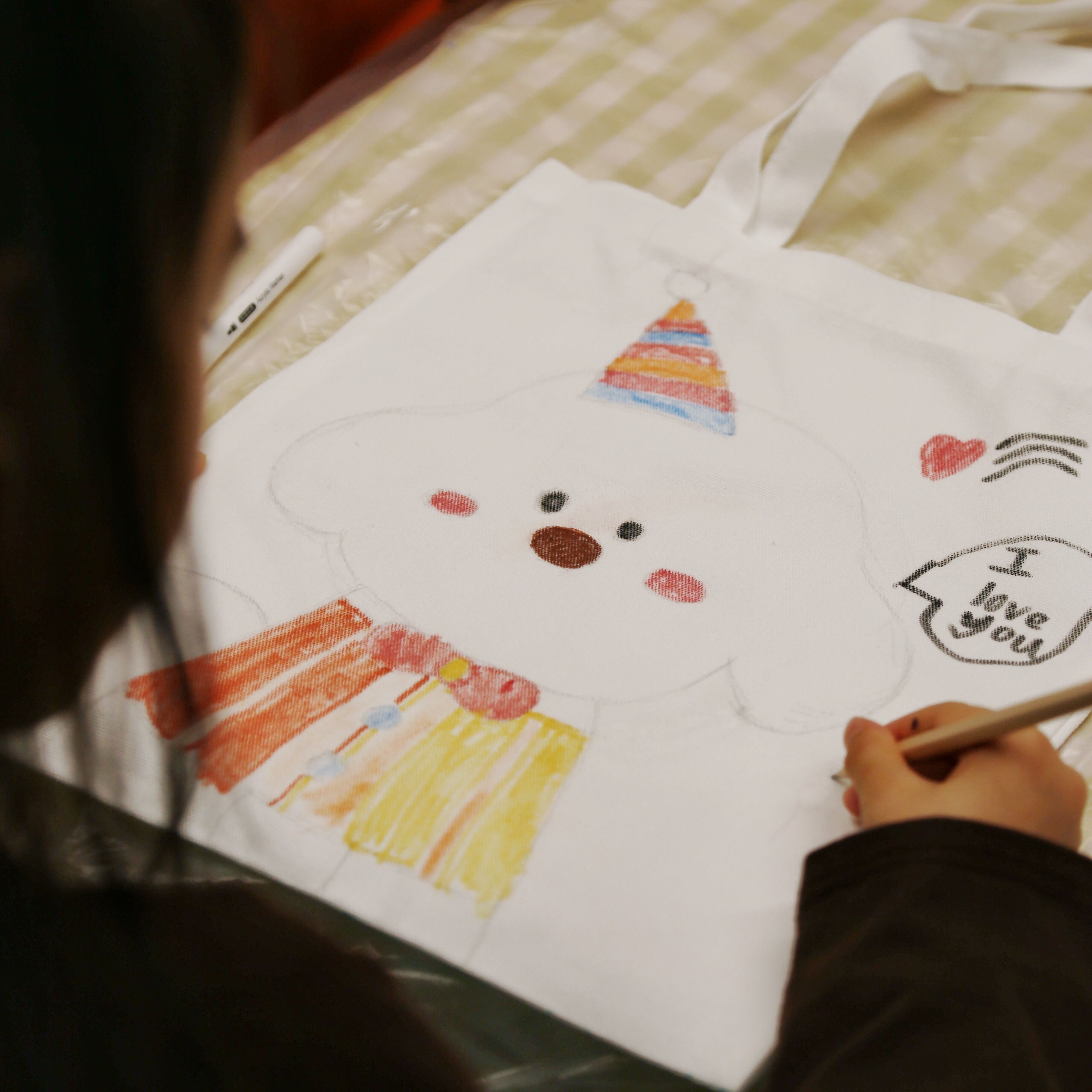 A person drawing on a white tote bag with a face design, including a hat and a heart with text.