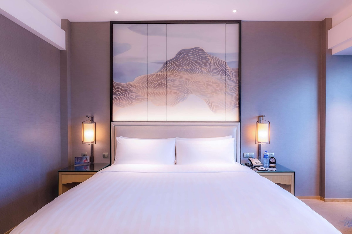 Ein modernes Hotelzimmer mit einem großen Bett, zwei Nachttischen mit Lampen und einem Wandbild, das eine Berglandschaft zeigt.