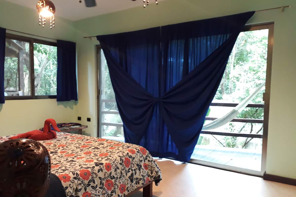 Ein Schlafzimmer mit einem Bett, einem Vorhangfenster, einem Deckenventilator und Blick ins Grüne.