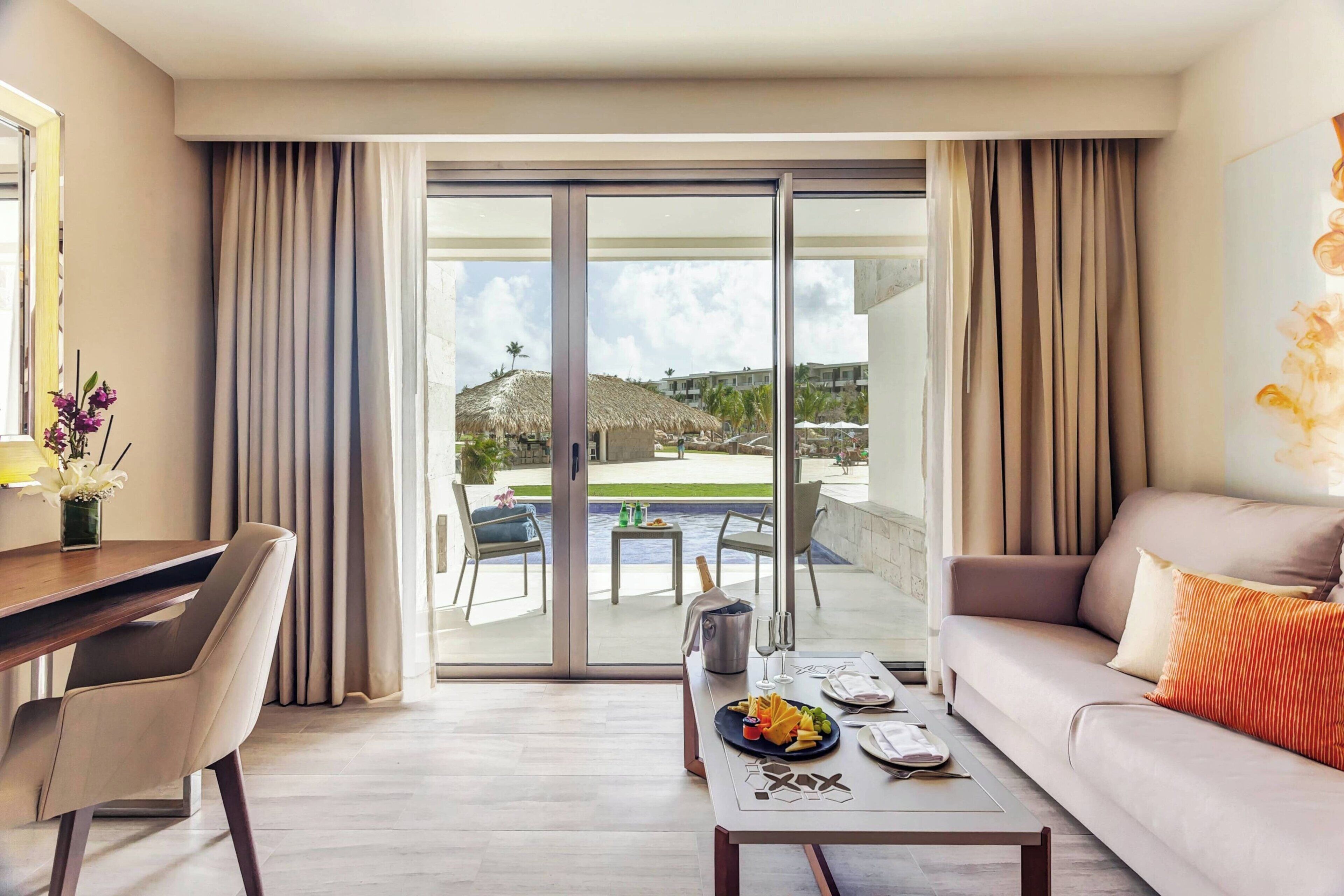 Una habitación de hotel moderna con puerta corrediza de vidrio que da a una zona de piscina, un sofá, una mesa de comedor y un escritorio.