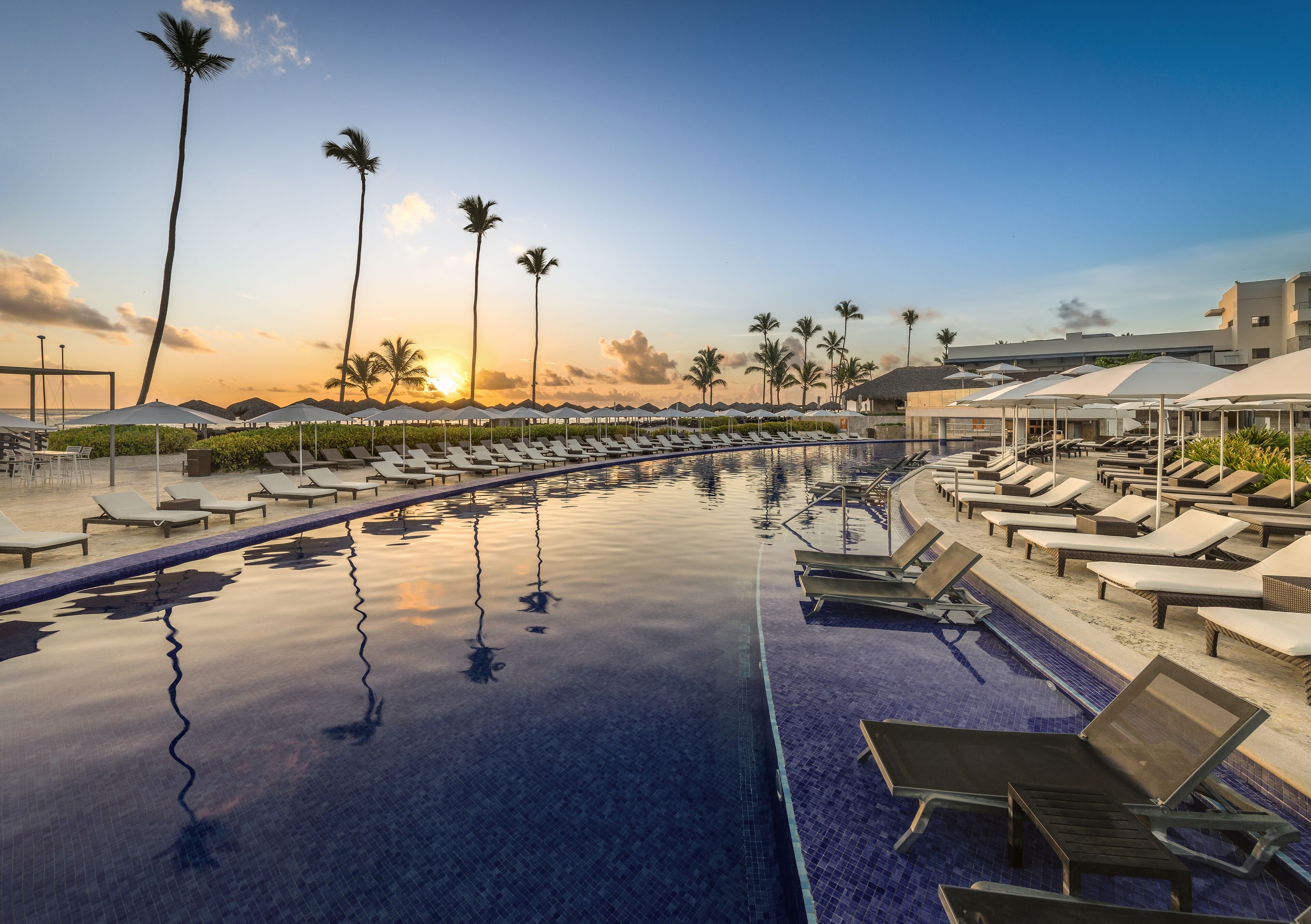 A poolside area with lounge chairs and umbrellas, palm trees, and a sunset view.