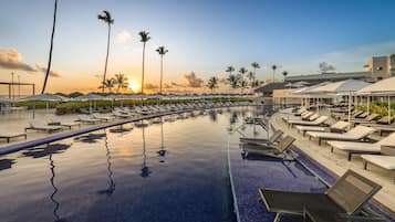Zona de piscina con sillones de descanso y sombrillas, palmeras y vistas al atardecer.
