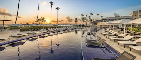 A poolside area with lounge chairs and umbrellas, palm trees, and a sunset view.