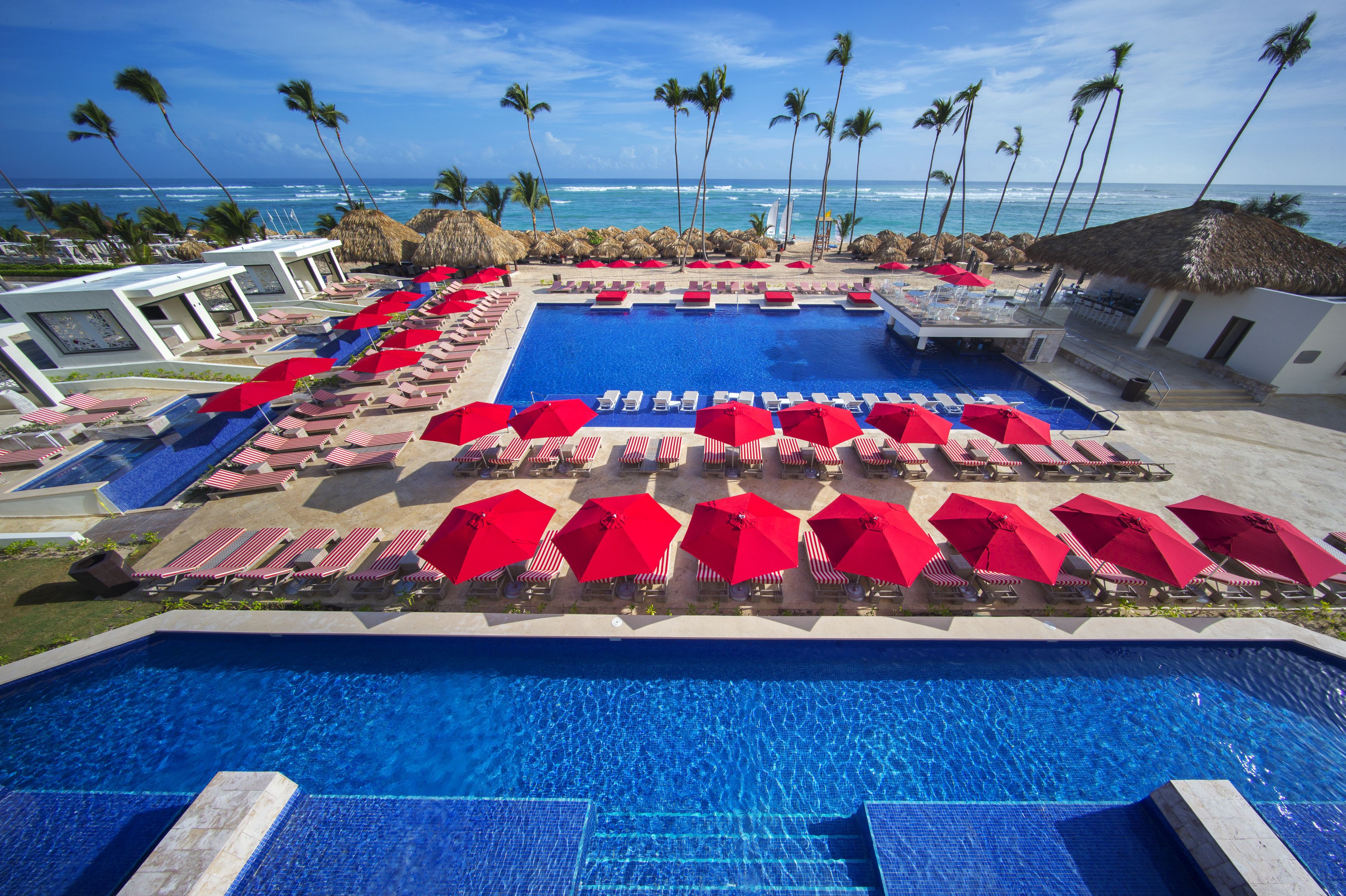 A resort pool area with multiple lounge chairs and umbrellas, a large central pool, and a view of the ocean.
