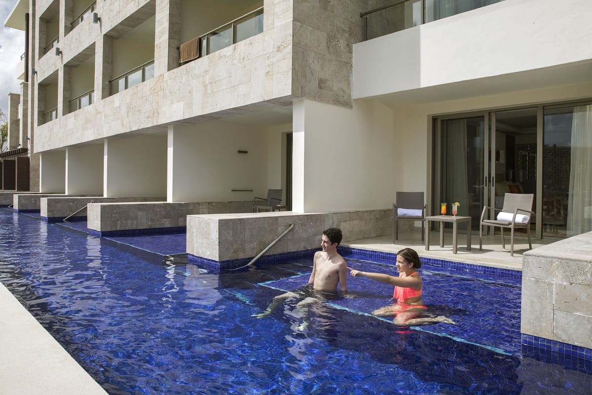 A swimming pool with a clear blue water feature, a modern building with balconies, and a seating area with a table and chairs.