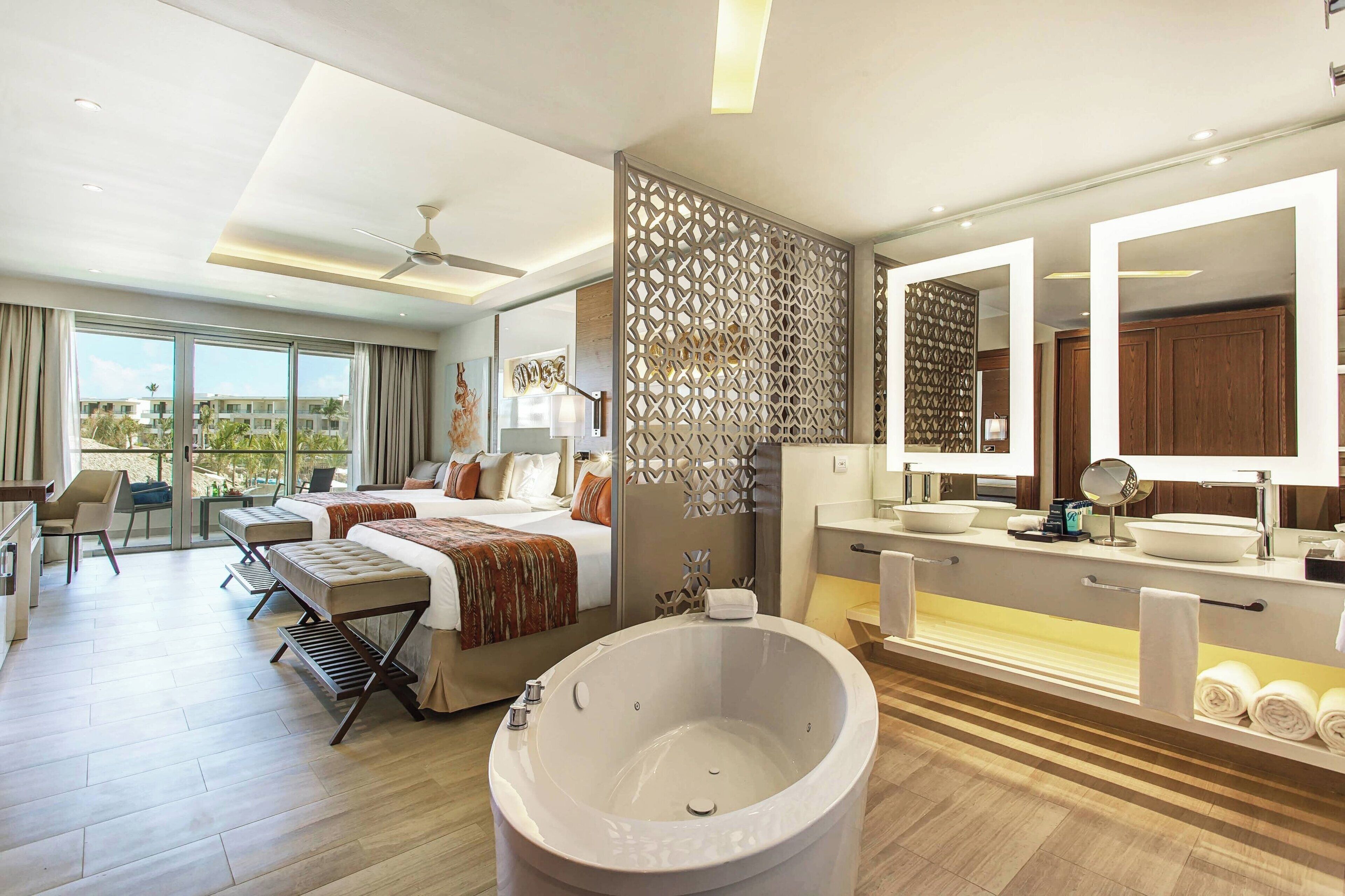 A modern hotel room with a large bed, a separate sitting area with a chair and table, a large bathtub, and a bathroom with a vanity and mirrors.
