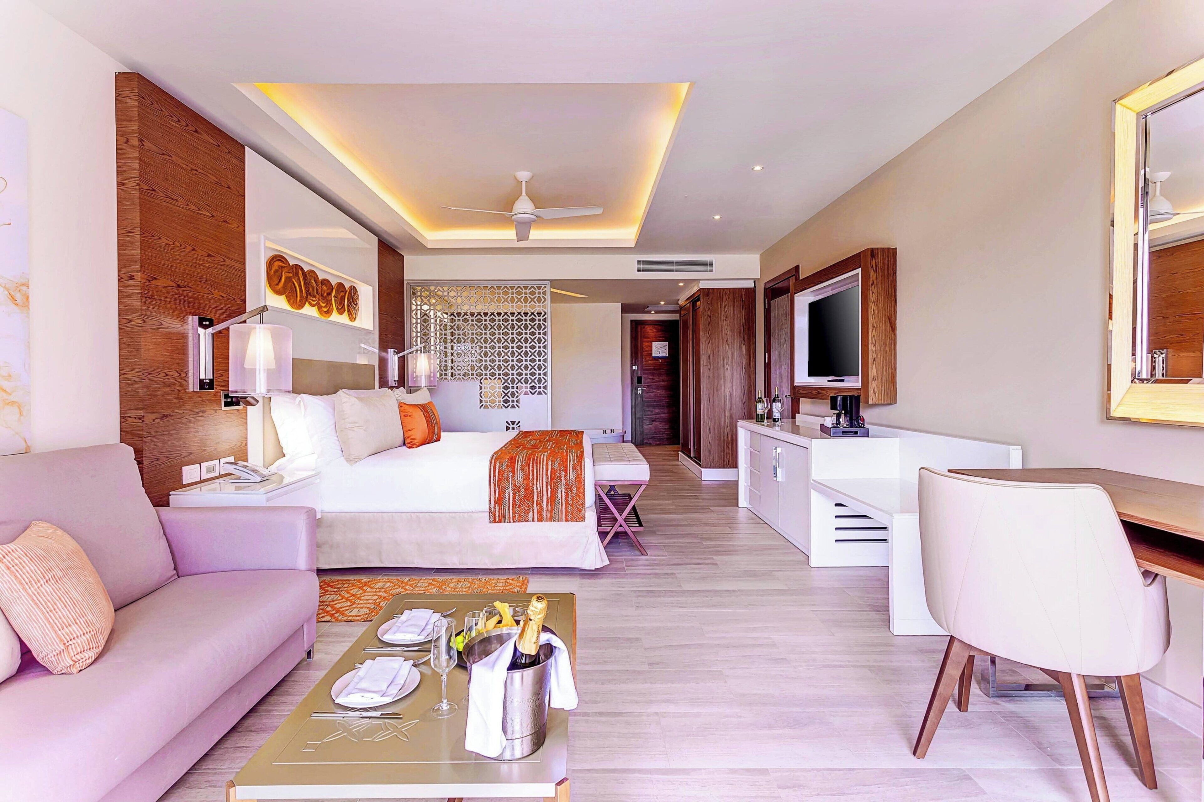 A modern hotel room with a bed, a sofa, a dining table with a bottle of champagne, a TV, and a desk.