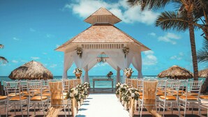 Allestimento per un matrimonio sulla spiaggia, con un gazebo in legno, sedie bianche e una navata bianca.